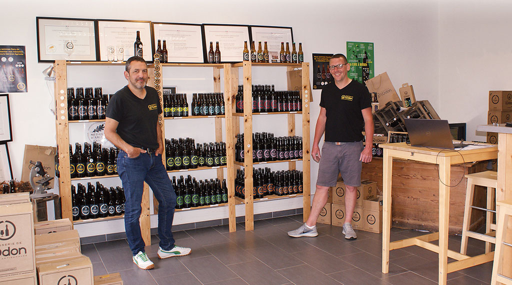 Deux hommes se tiennent dans une brasserie, entourés d'étagères remplies de bouteilles de bière et de matériel de brassage.