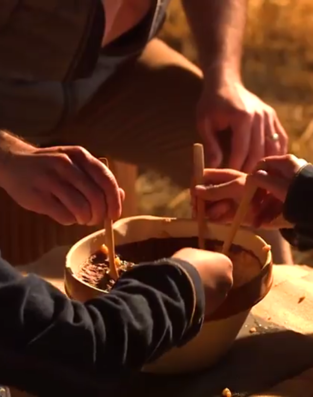 Des mains utilisant des bâtonnets en bois pour mélanger un mélange dans un grand bol, entourées d'un cadre rustique.